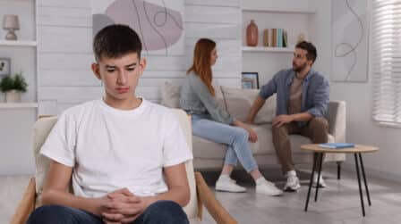 Nastolatek w białej koszulce siedzi na pierwszym planie ze spuszczoną głową i zmartwionym wyrazem twarzy. W tle dwoje dorosłych rozmawia na kanapie, co sugeruje trudną sytuację rodzinną, podczas której rodzice mają trudności dotyczące tego, jak rozmawiać z nastolatkiem o rozwodzie.
